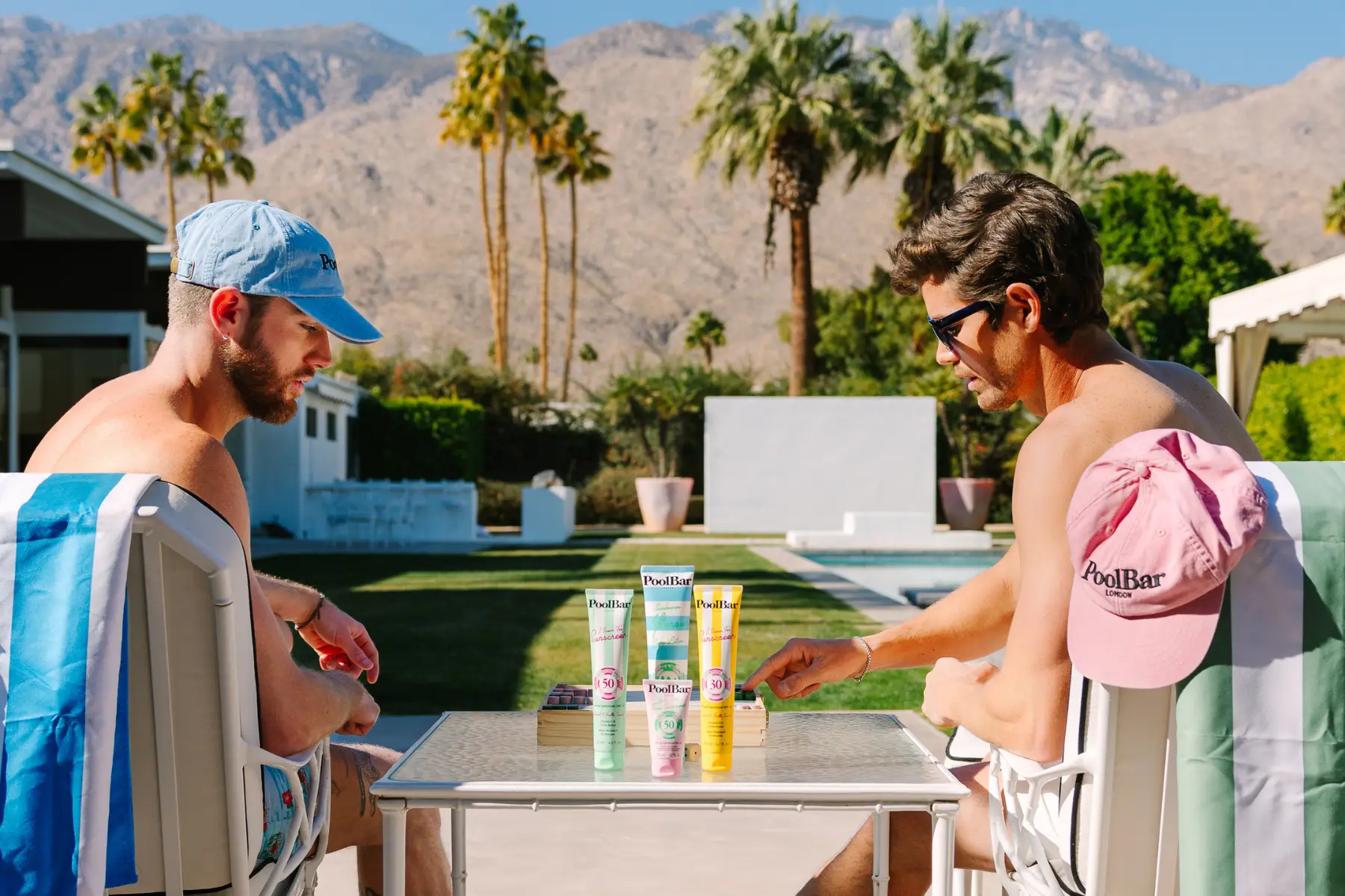 Two men sat in the sun playing chess with PoolBar sunscreen on the table