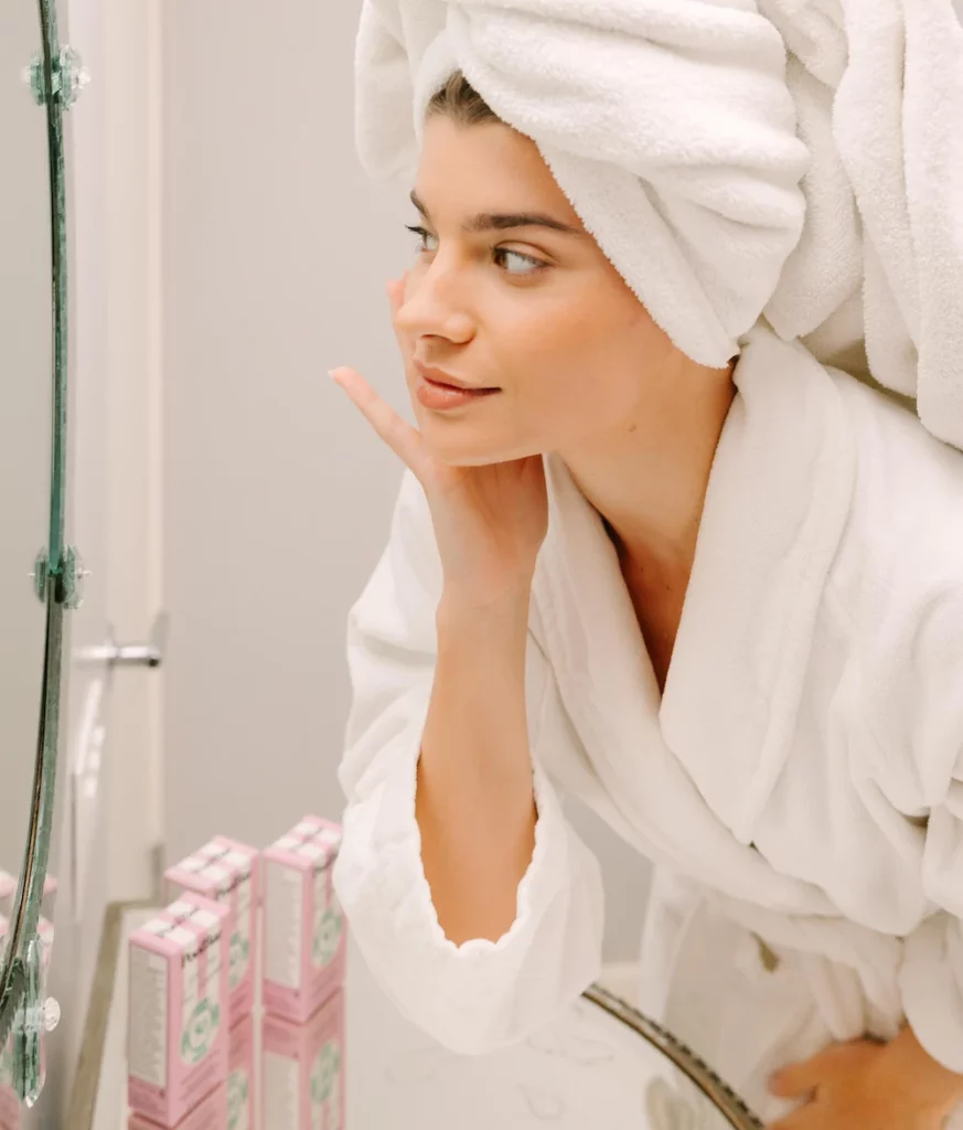 woman in bath robe and hair towel applying face sunscreen in the bathroom mirror